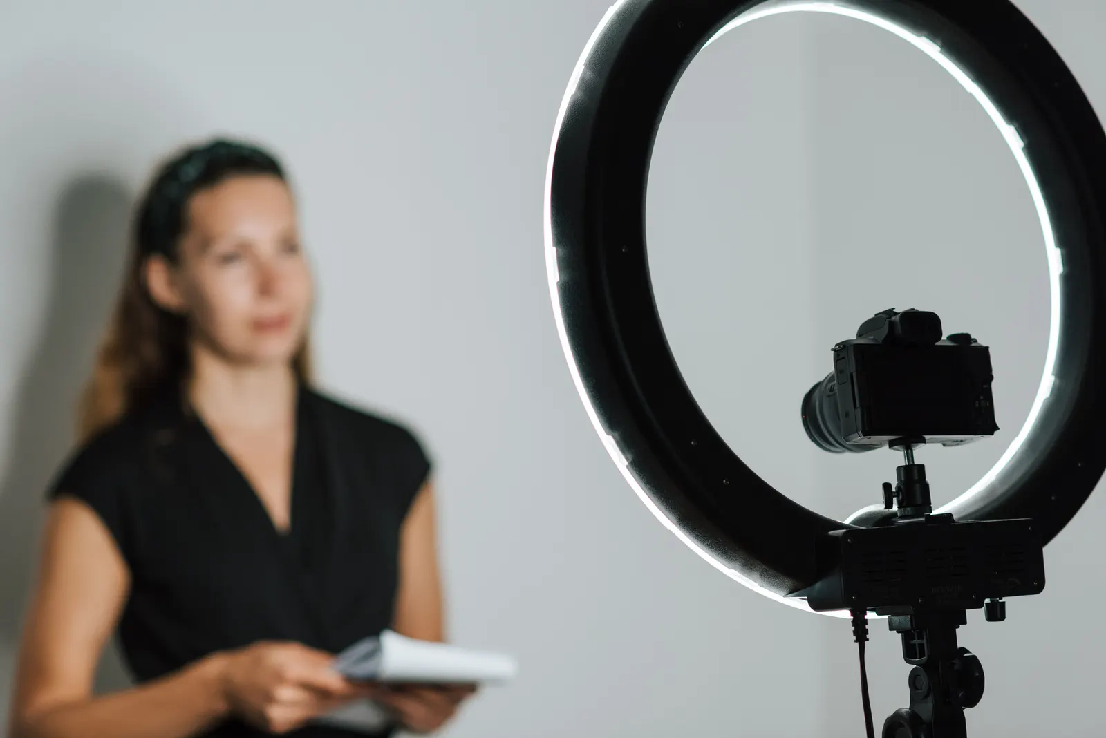 A ring light and a camera on a tripod, with a woman out of focus in the background holding some papers.